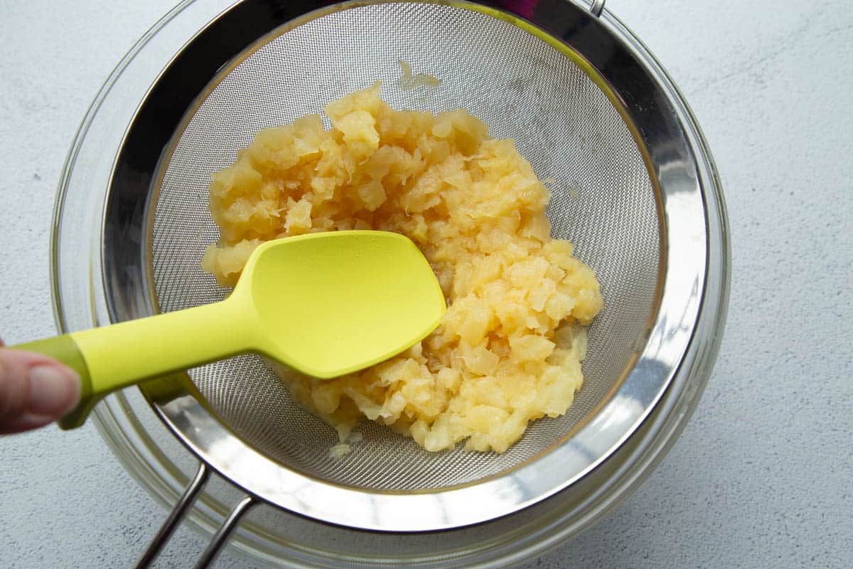 a yellow spatula pushing on a colander full of crushed pineapple.