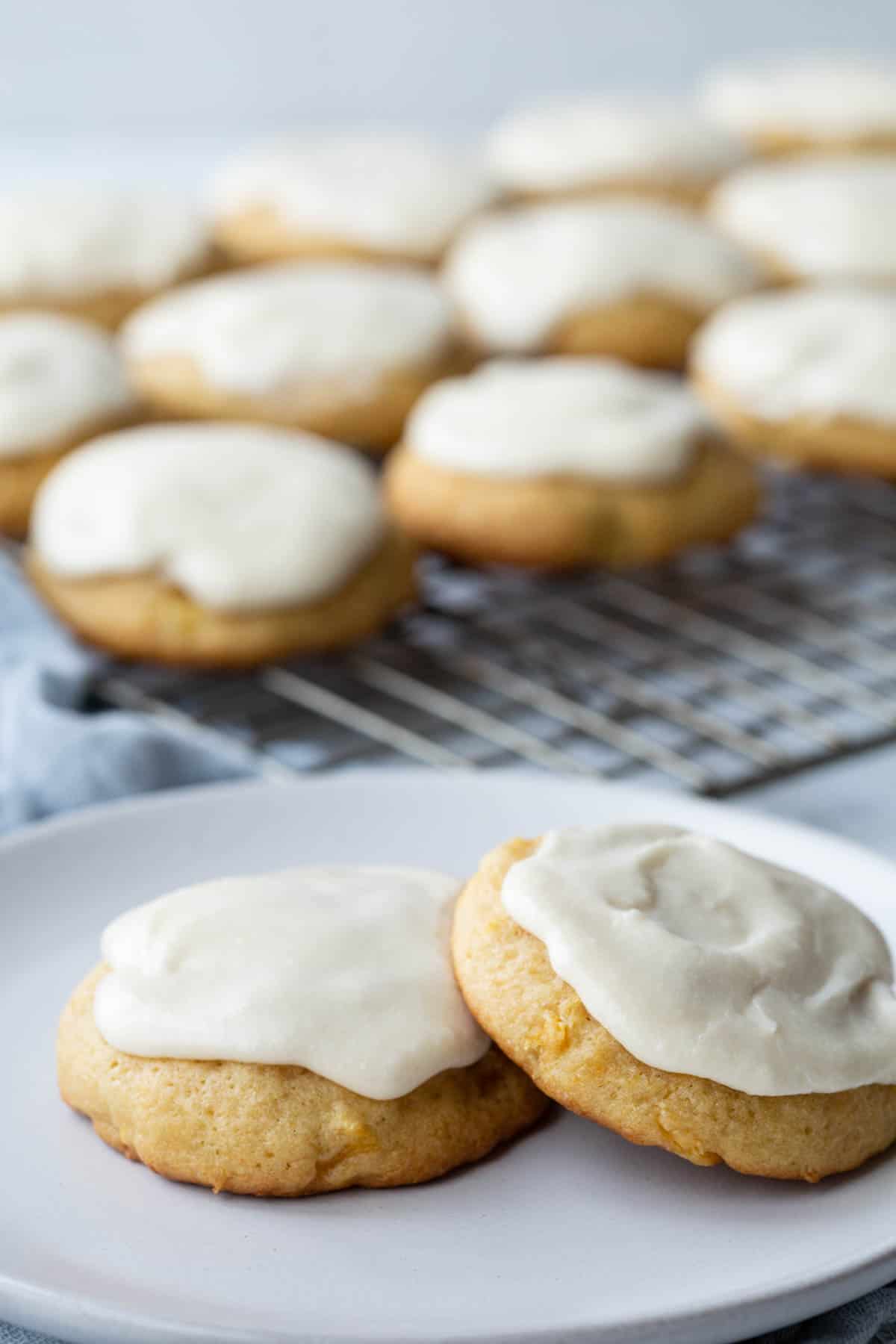 two iced pineapple cookies on a white plate, in front of a wire rack filled with more pineapple cookies.