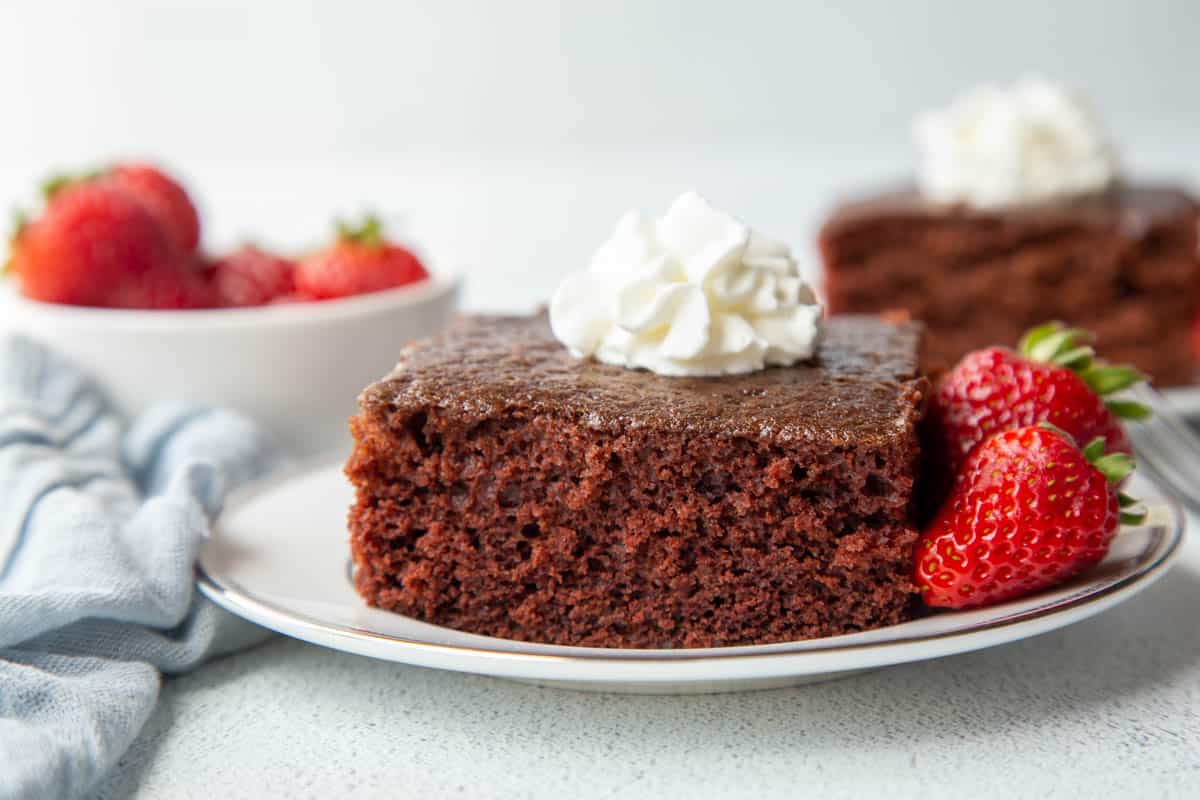 slice of wacky cake on a plate, surrounded by strawberries and topped with whipped cream.