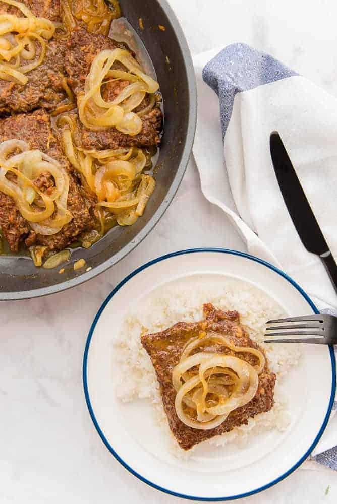 cube steak smothered in onions both in a skillet with a single serving on a white plate.