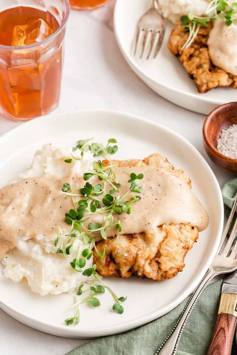 chicken fried steak topped with a creamy gravy on a white plate with an herb garnish.