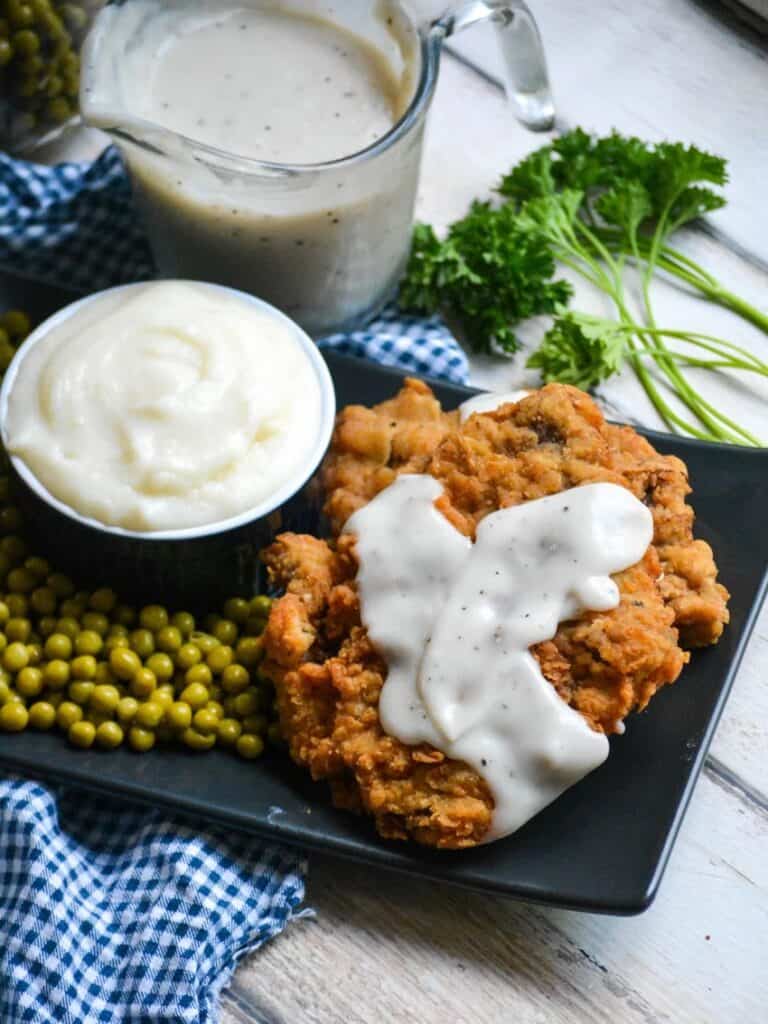 chicken fried steak topped with white gravy on a black platter with peas and more white gravy.