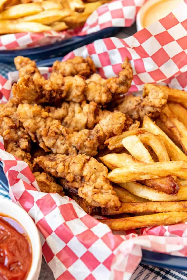 fried steak strips in a red and white paper lined basket next to french fries.