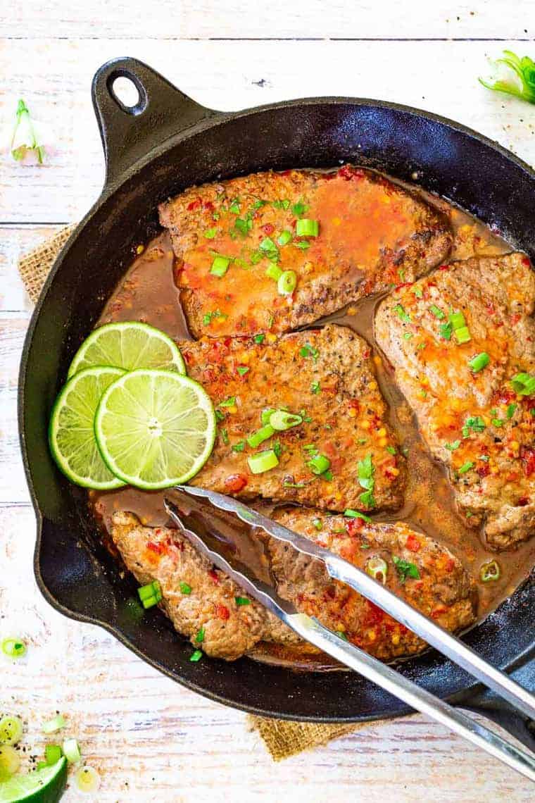 cube steaks in a skillet in a chili sauce, along with sliced limes.