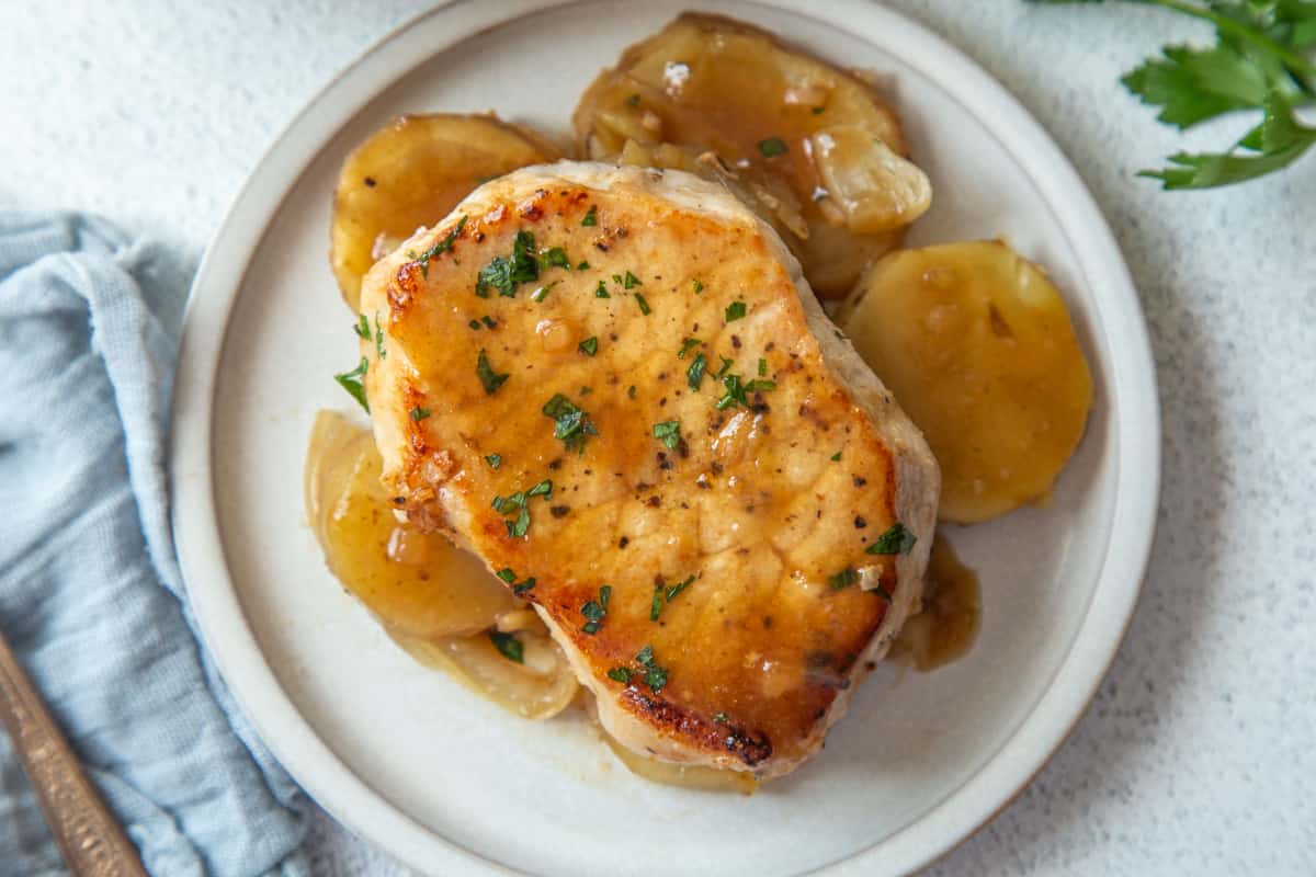 pork chop covered in light brown sauce on top of sliced potatoes on a white plate.