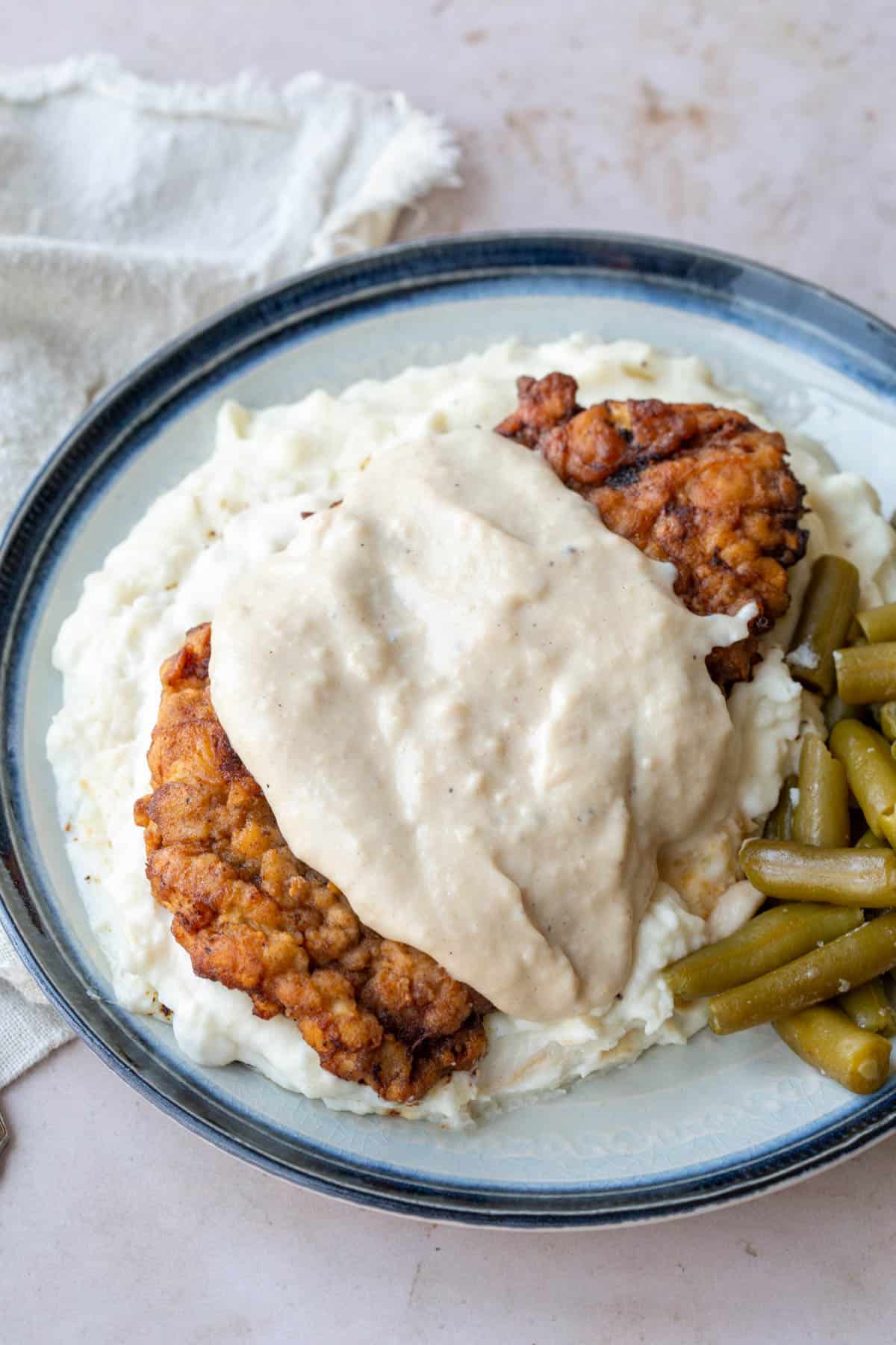 chicken fried steak topped with white gravy on a plate with mashed potatoes and green beans.