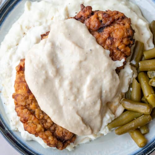 piece of chicken fried steak with white gravy on top of mashed potatoes with green beans on the side.