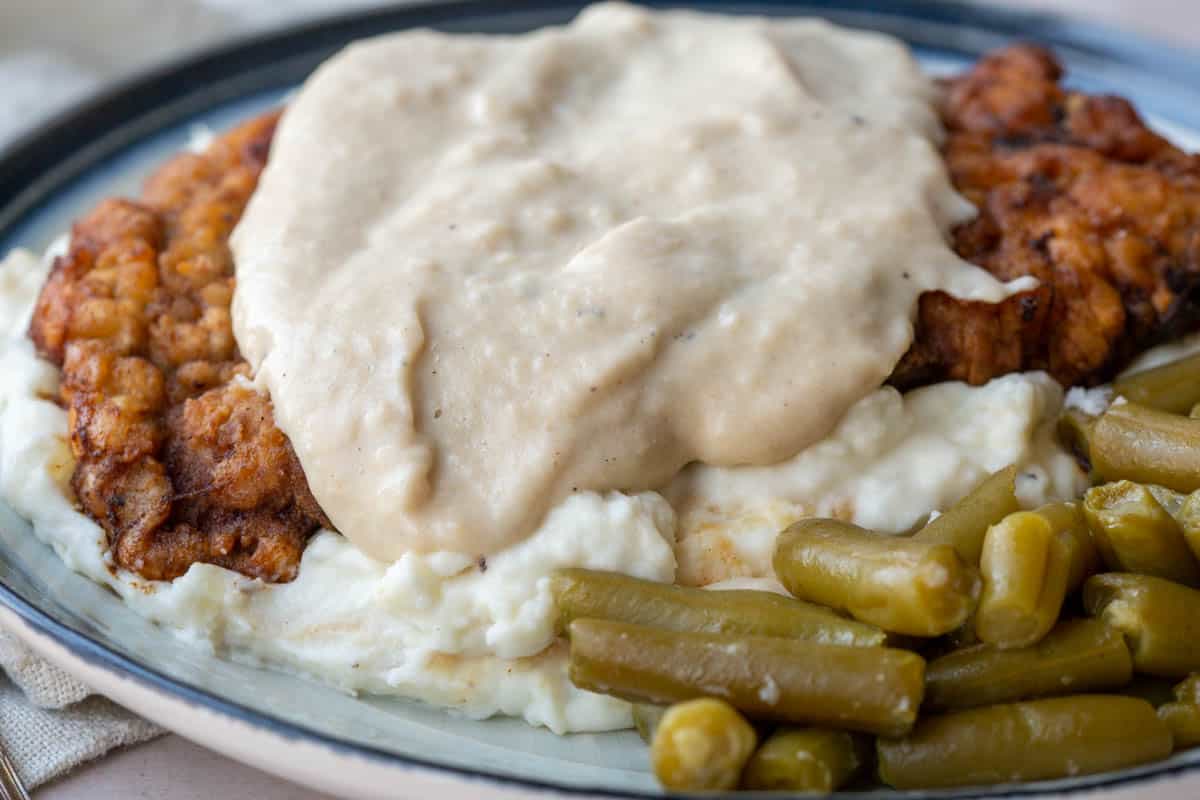 chicken fried steak covered in white gravy on a plate with mashed potatoes and green beans.
