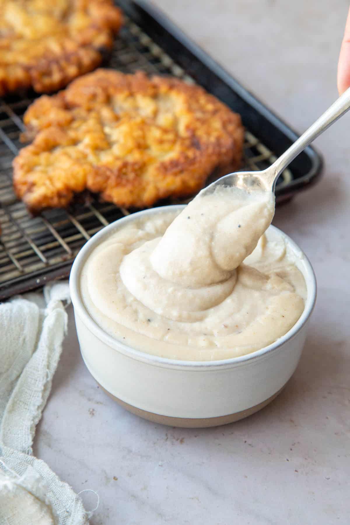a spoon holding a spoonful of white gravy in front of a sheet pan of chicken fried steak.