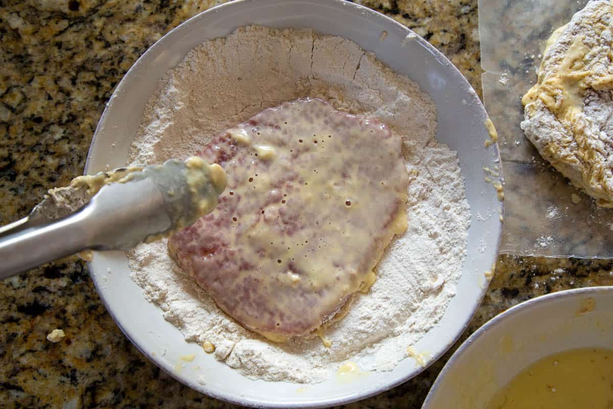 tongs dipping a cube steak coated in egg into a flour mixture in a shallow white dish.