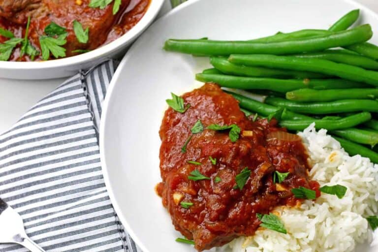 swiss steak covered in tomato gravy with mashed potatoes and green beans on a white plate.