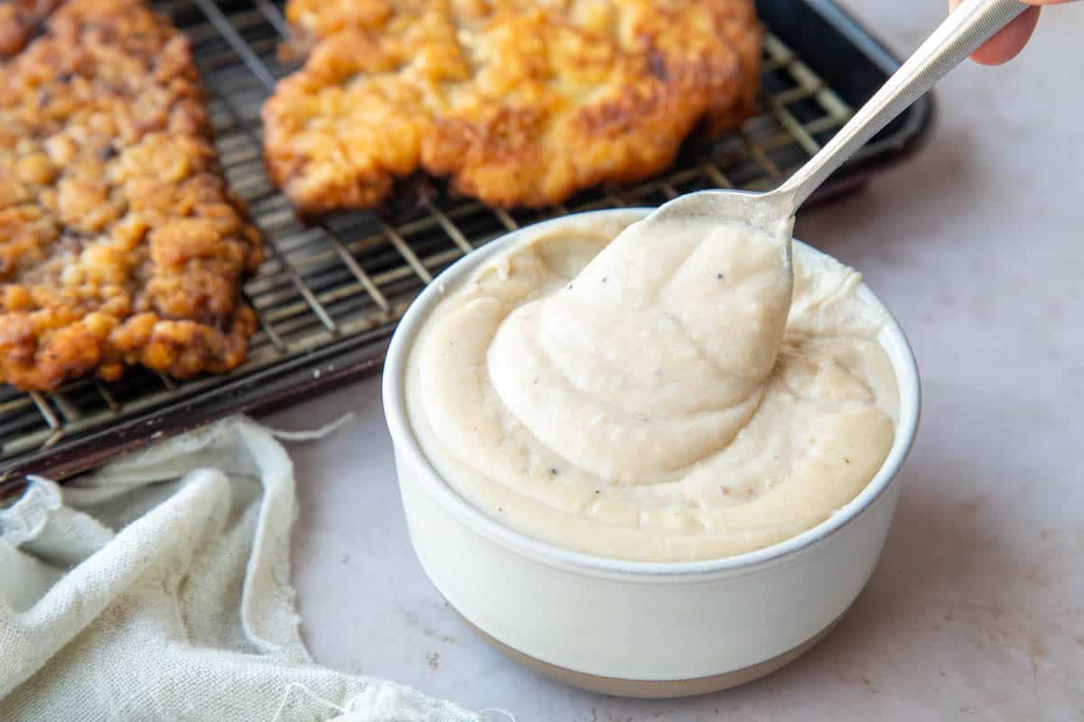 spoonful of white gravy above a dish of more white gravy, with chicken fried steak in the background.