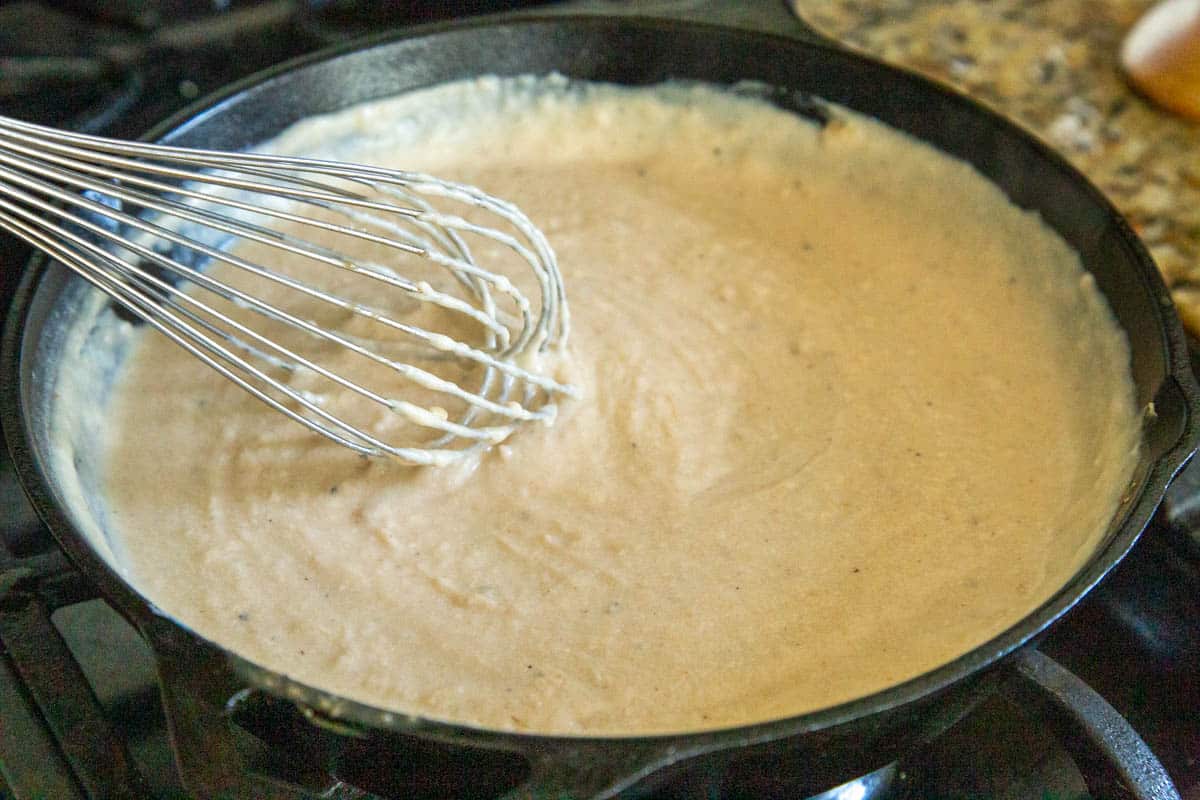 whisk stirring white gravy in a cast iron skillet.