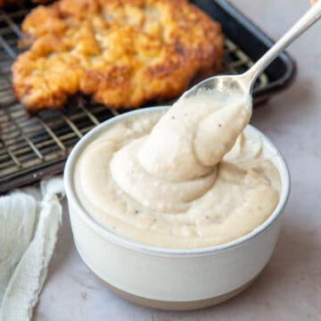 spoon holding a spoonful of easy white gravy with black pepper flecks in front of a pan of chicken fried steak.