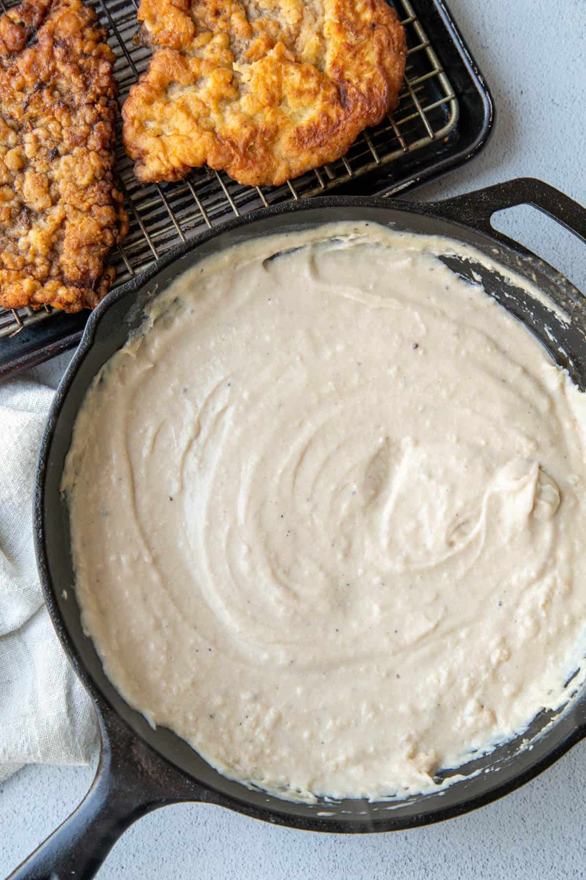 white gravy in a cast iron skillet, next to chicken fried steak on a sheet pan.