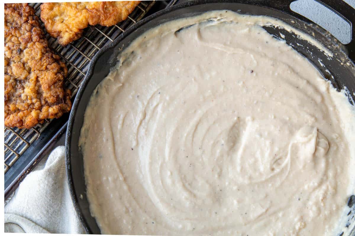 white gravy in a cast iron skillet next to a sheet pan filled with chicken fried steak.