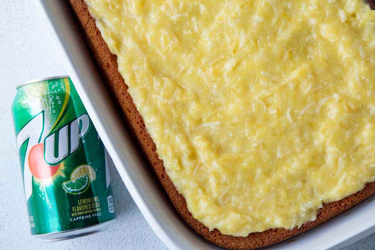 whole 7up cake in a white baking dish, topped with a yellow pineapple frosting, next to a can of 7up soda.