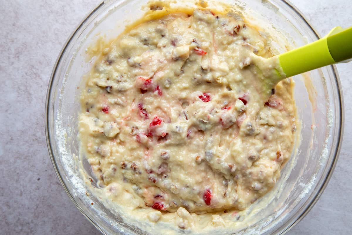 batter with cherries and pecans in a glass bowl with a yellow spatula.