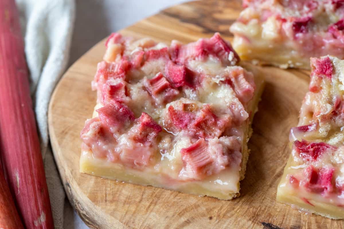 rhubarb bars on a wooden board, next to several stalks of fresh rhubarb.