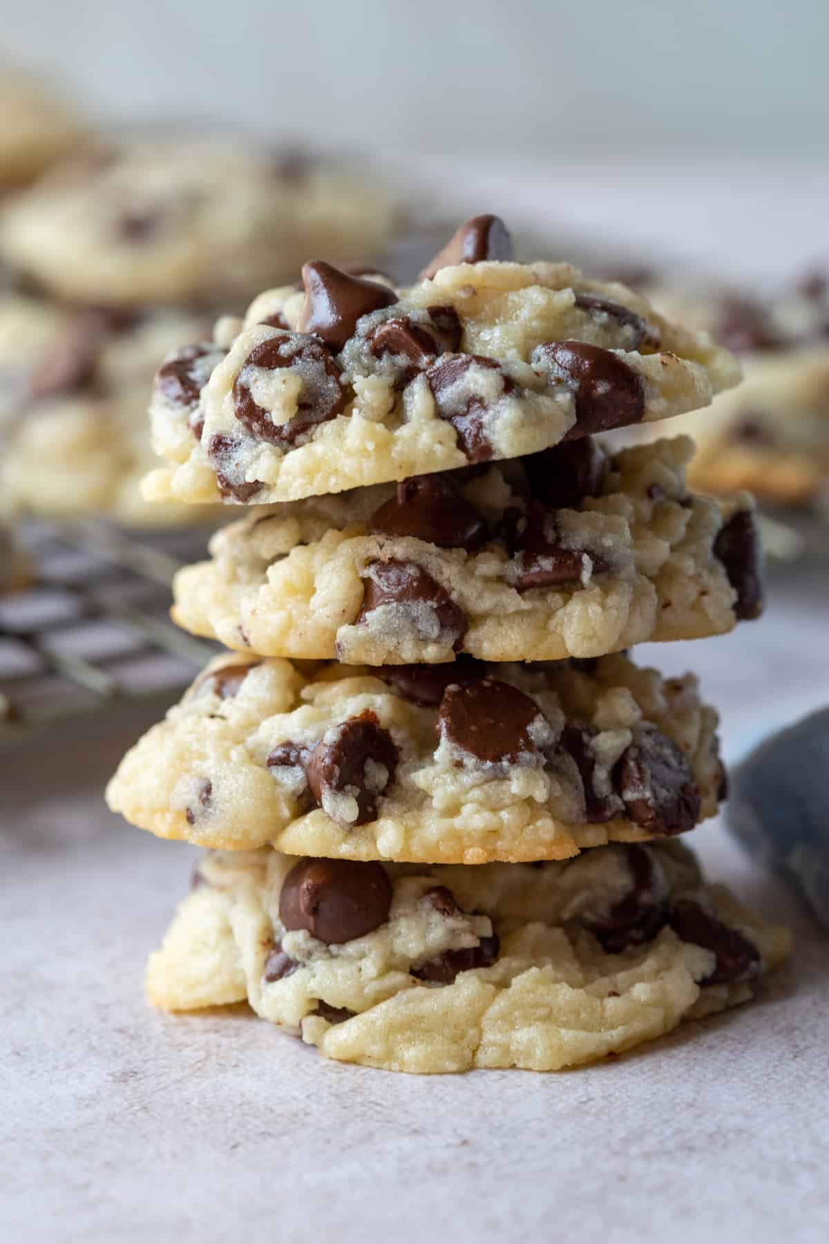 stack of cream cheese chocolate chip cookies with more cookies on a wire rack in the background.