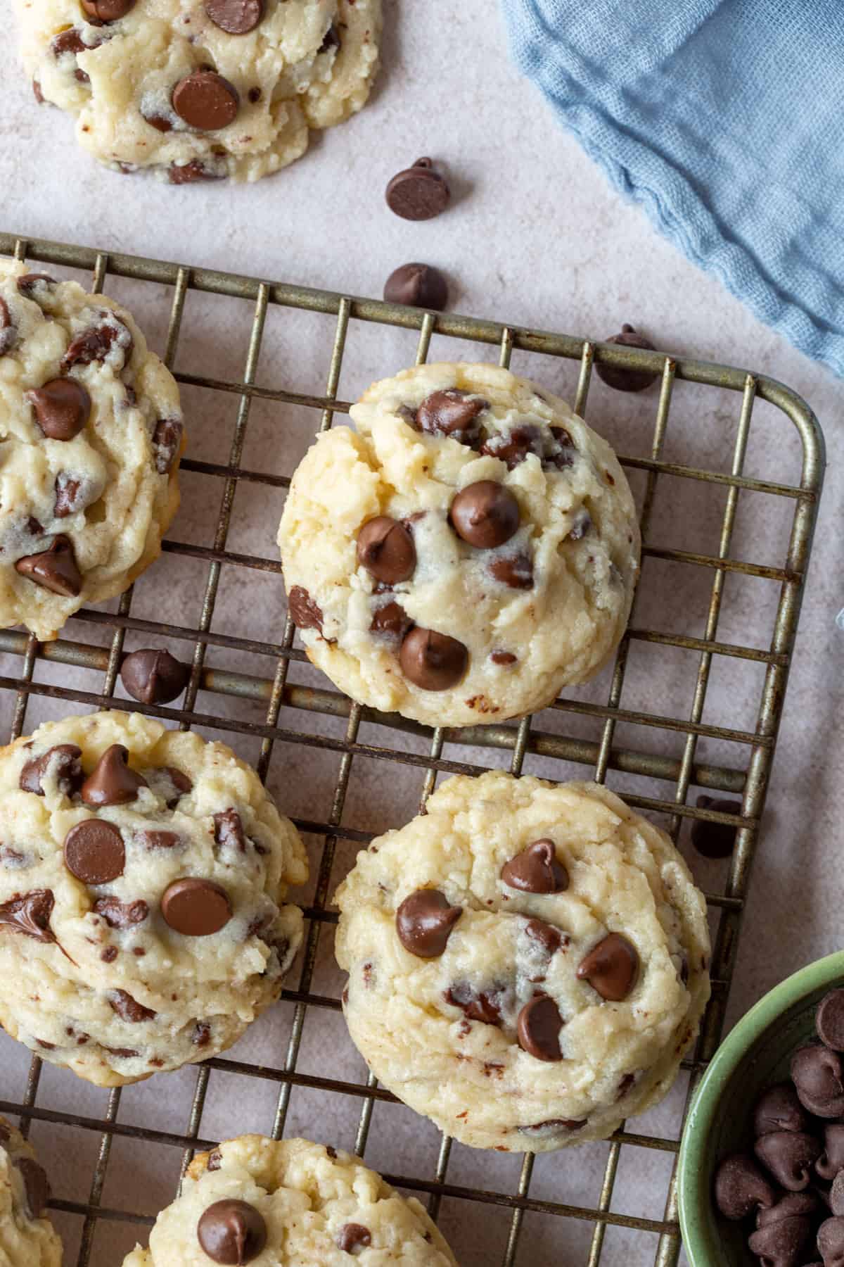 cream cheese chocolate chip cookies on a wire rack next to a few chocolate chips and a blue tea towel.