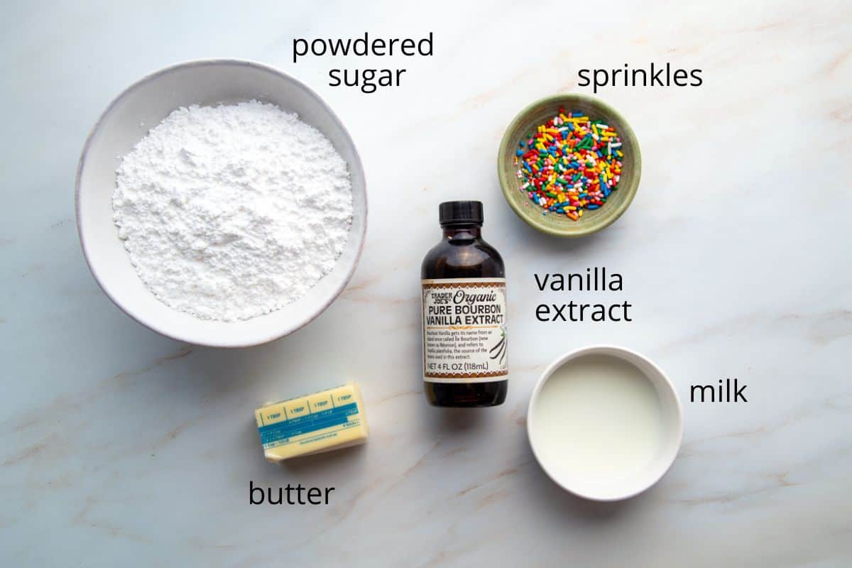 powdered sugar, vanilla, butter, milk, and sprinkles on a white marble countertop.