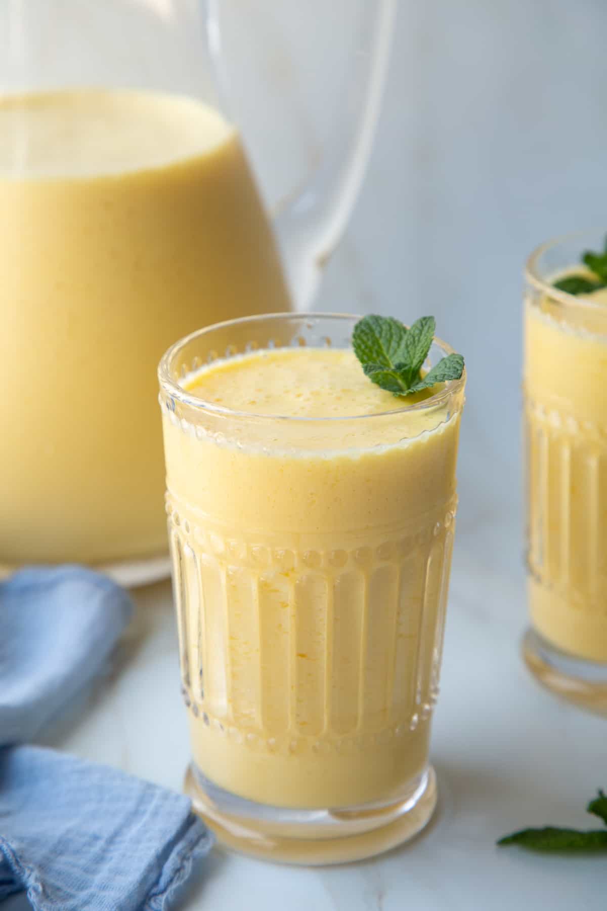 homemade orange julius in two decorative glasses, with a pitcher of more orange julius drink behind them.