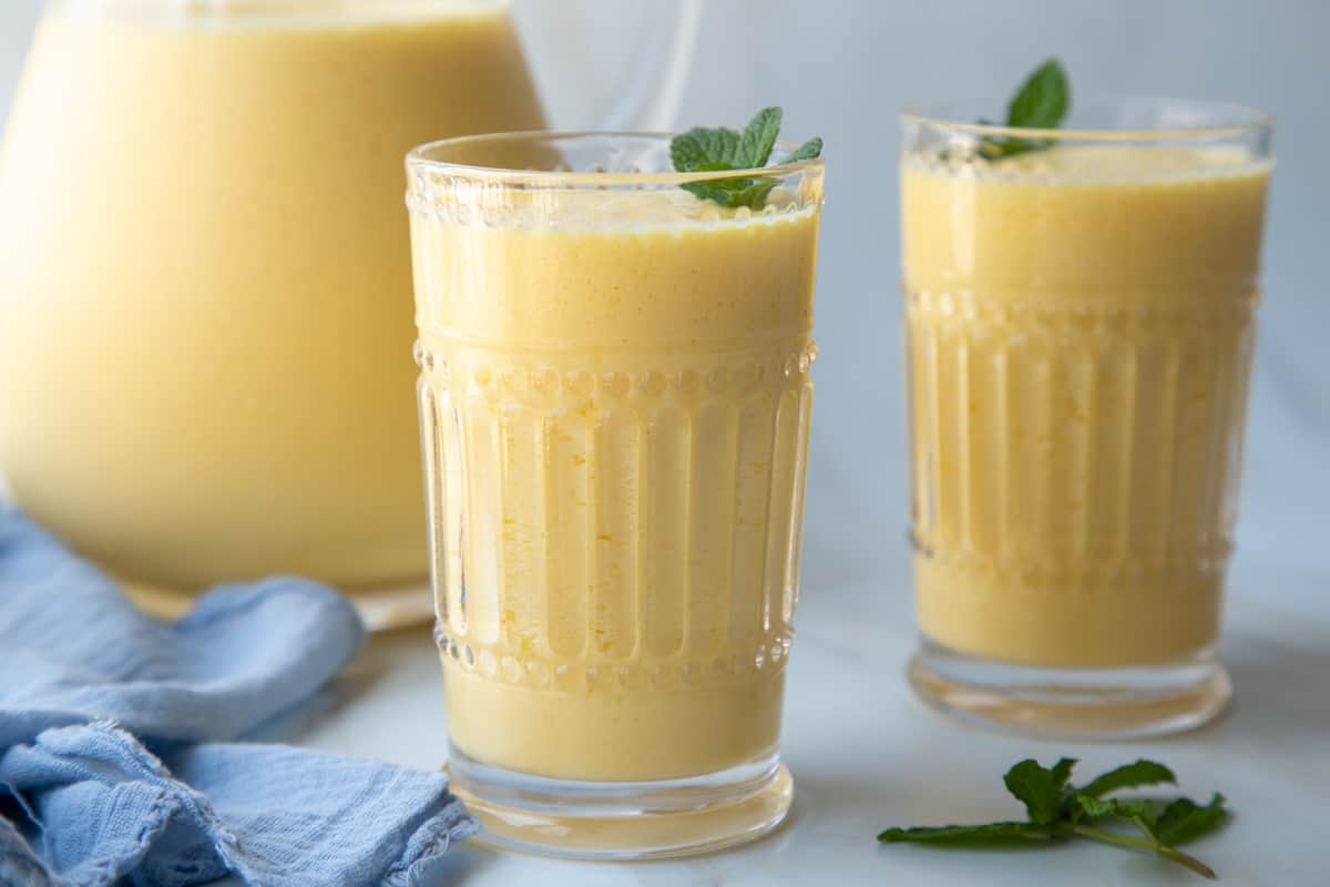 orange julius in two decorative glasses and also in a glass pitcher, next to a blue tea towel. 