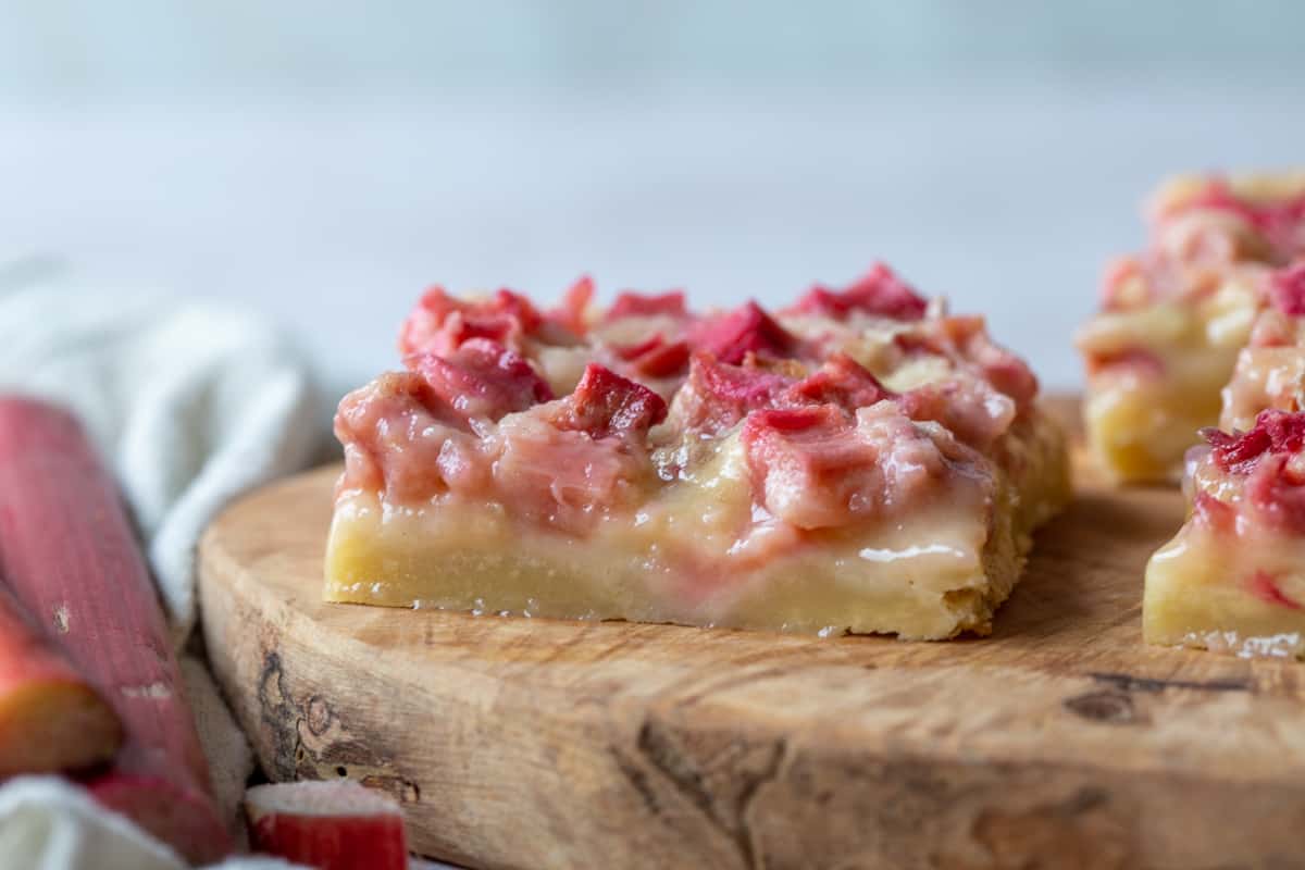 rhubarb bars on a shortbread crust on a wooden board, next to stalks of fresh rhubarb.