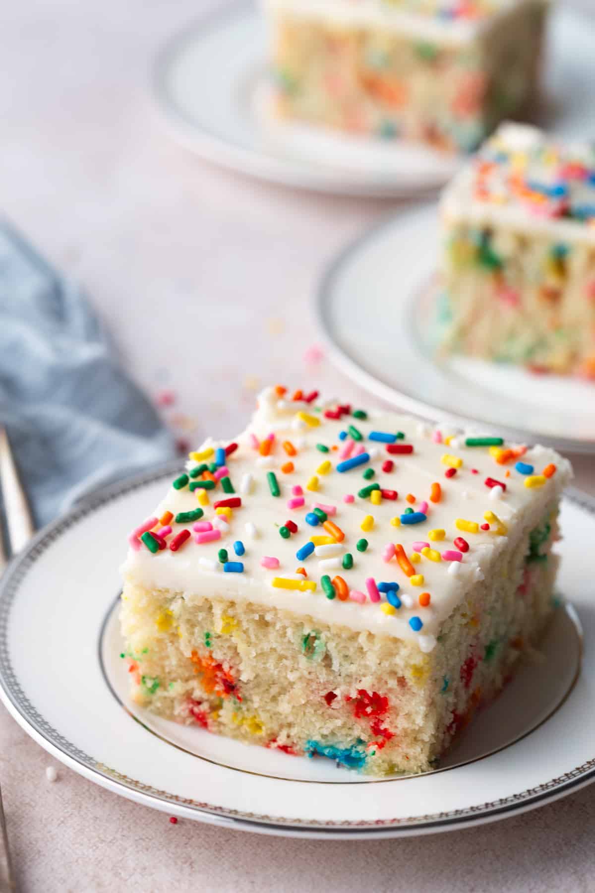 three pieces of vanilla wacky cake with sprinkles and frosting on white plates with a silver rim.