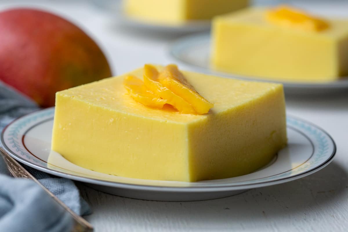 square slice of creamy mango salad garnished with fresh mango slices on a white plate with a blue rim.