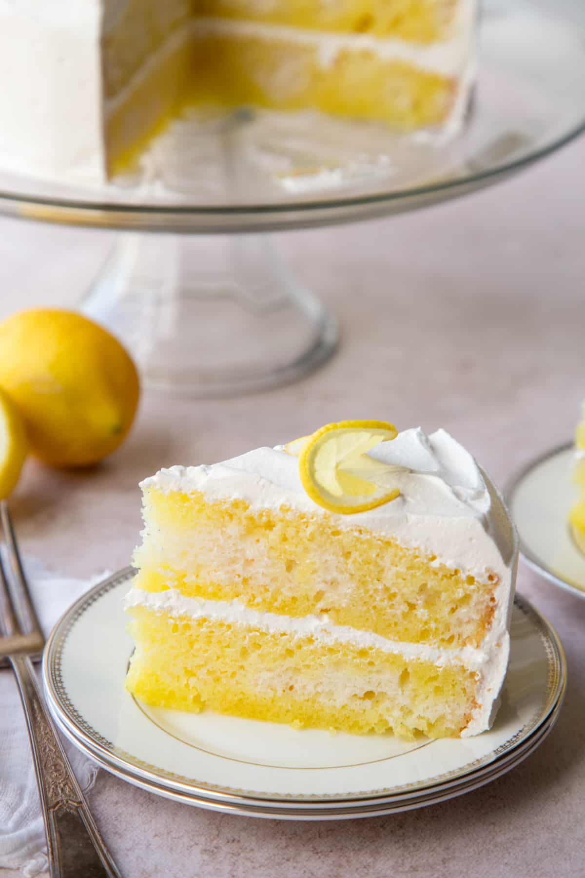 slice of lemon poke cake on a white plate next to a whole cake.