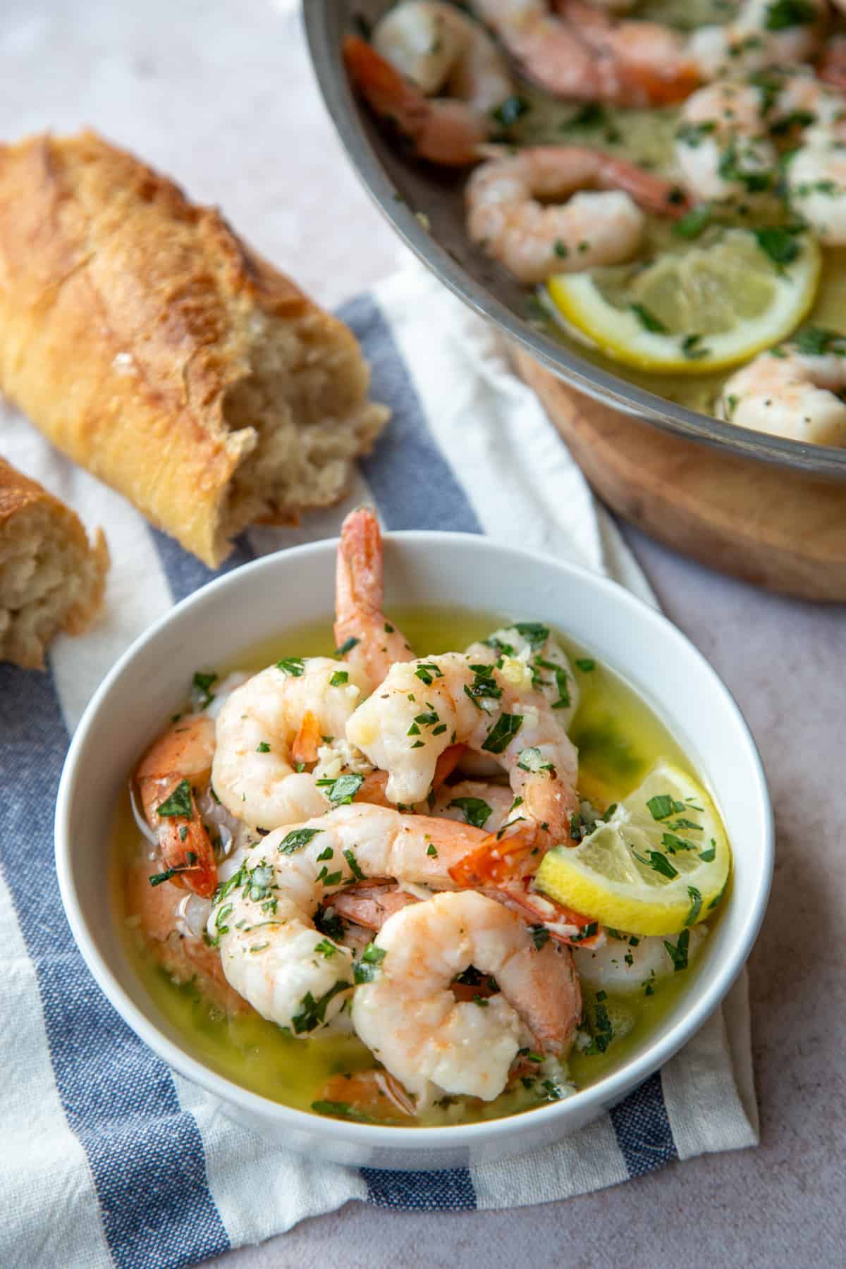 small dish of shrimp scampi next to a baguette on a blue and white tea towel.