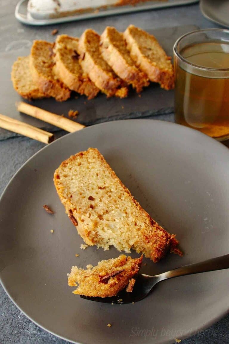 A slice of Cinnamon Tea Cake with Pears on a grey plate with tea, cinnamon and more cake in the background.