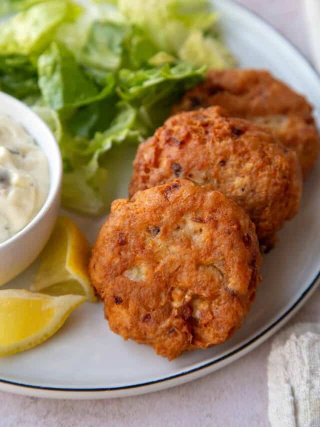 Top view of 2 fried Salmon Patties on a dinner plate next to Tartar Sauce and lemon wedges.