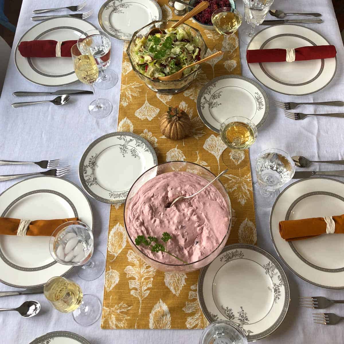 dinner table with a gold table runner, a pink jello salad in a bowl, plates, and orange napkins.