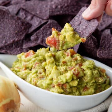 Close up of a hand lifting a blue corn tortilla chip from a bowl with Rotel Guacamole on it.