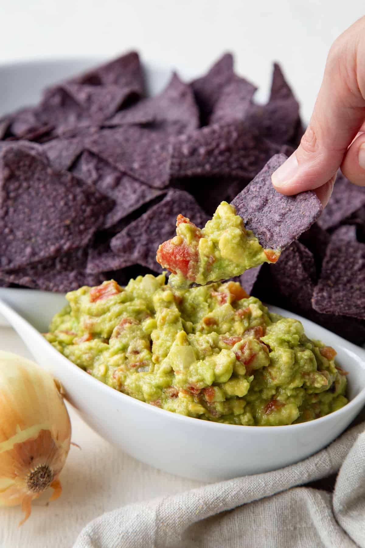 Hand lifting a blue corn tortilla chip from a bowl with Rotel Guacamole on it.