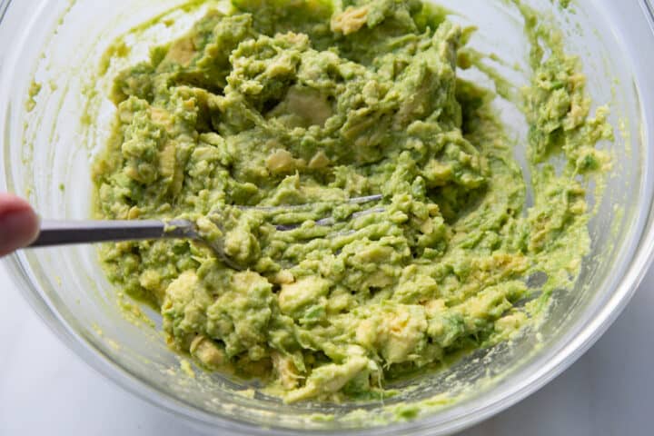 Fork mashing avocados in a clear glass mixing bowl for Rotel Guacamole.