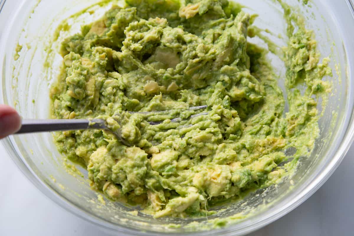 Fork mashing avocados in a clear glass mixing bowl.