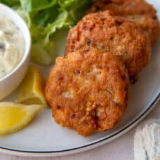 2 fried Salmon Patties on a dinner plate next to Tartar Sauce and lemon wedges.