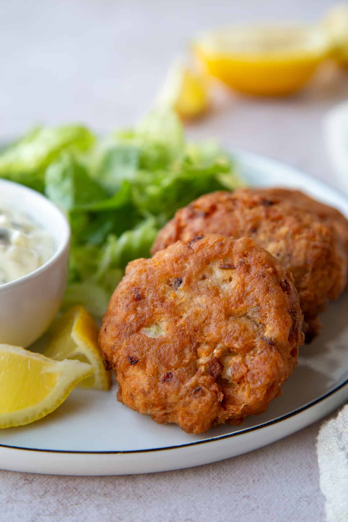 2 fried Salmon Patties on a dinner plate next to Tartar Sauce and lemon wedges.