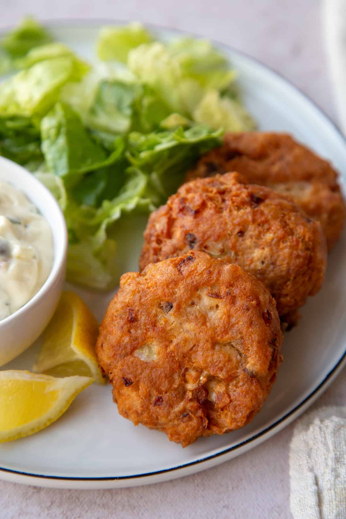 Top view of 2 fried Salmon Patties on a dinner plate next to Tartar Sauce and lemon wedges.