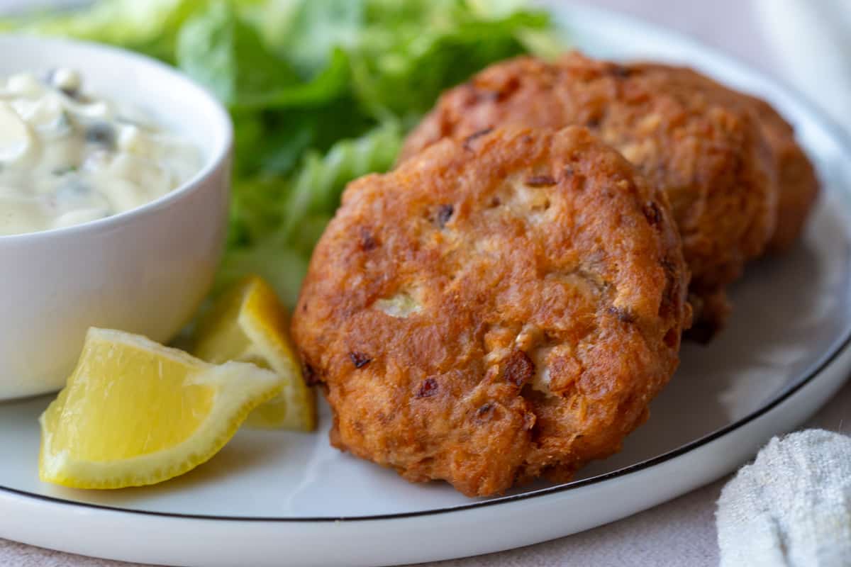 Closer view of 2 fried Salmon Patties on a dinner plate next to Tartar Sauce and lemon wedges.