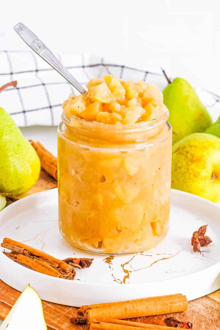 Side view of Pear Compote in a glass jar on a white plate next to pears and cinnamon sticks.