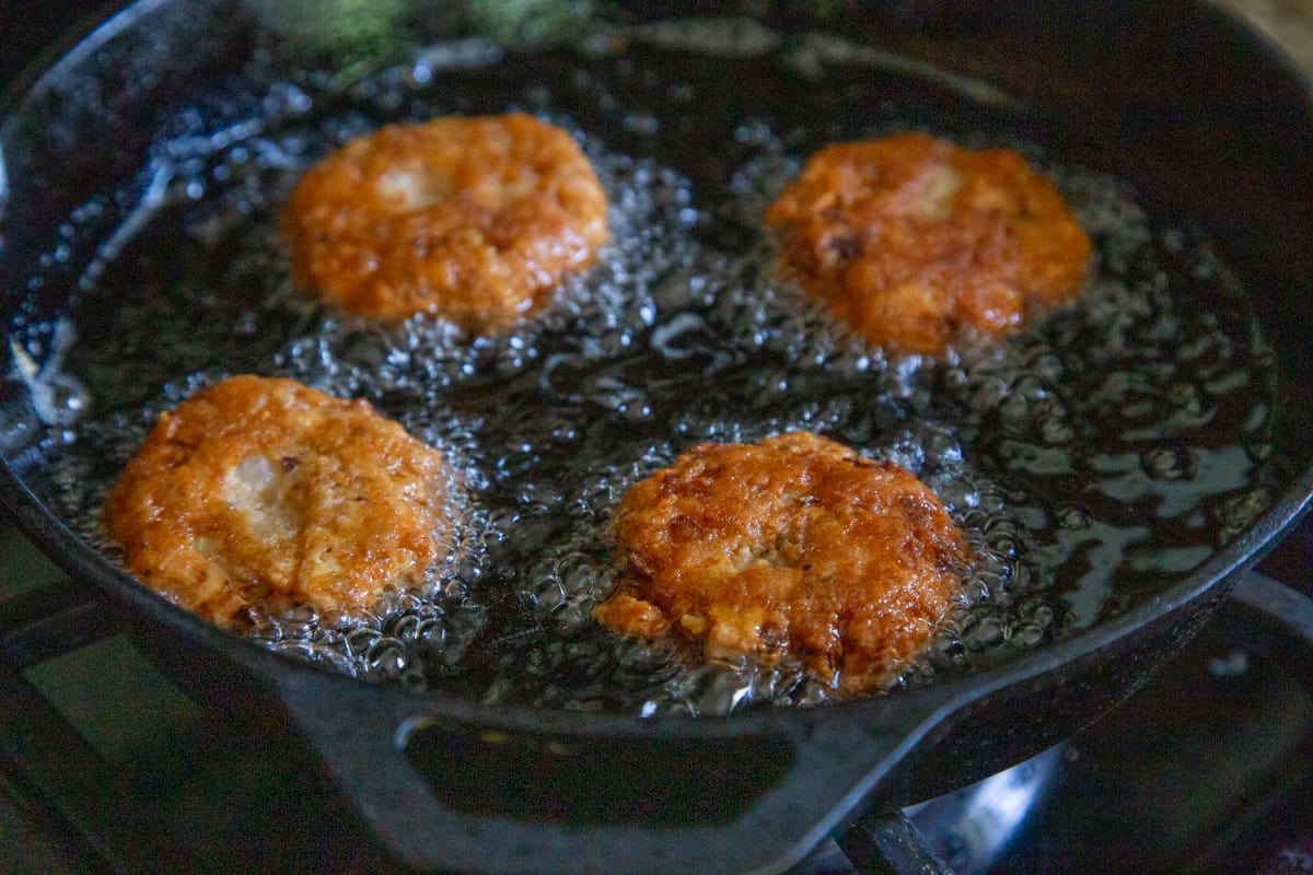 4 Salmon Cakes frying in a cast iron skillet.