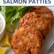 two fried Salmon Patties on a dinner plate next to Tartar Sauce and lemon wedges.