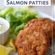 2 fried Salmon Patties on a dinner plate next to Tartar Sauce and lemon wedges.