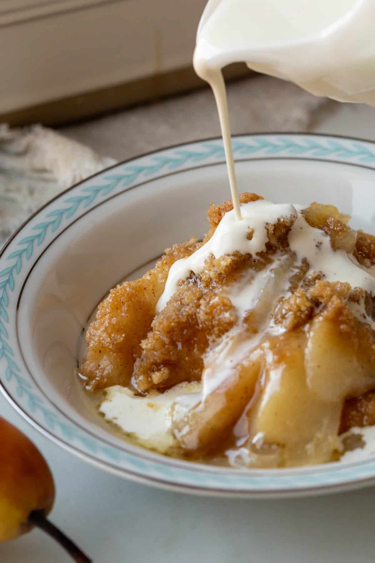 A small pitcher of cream being drizzled over a serving of Pear Crumble.
