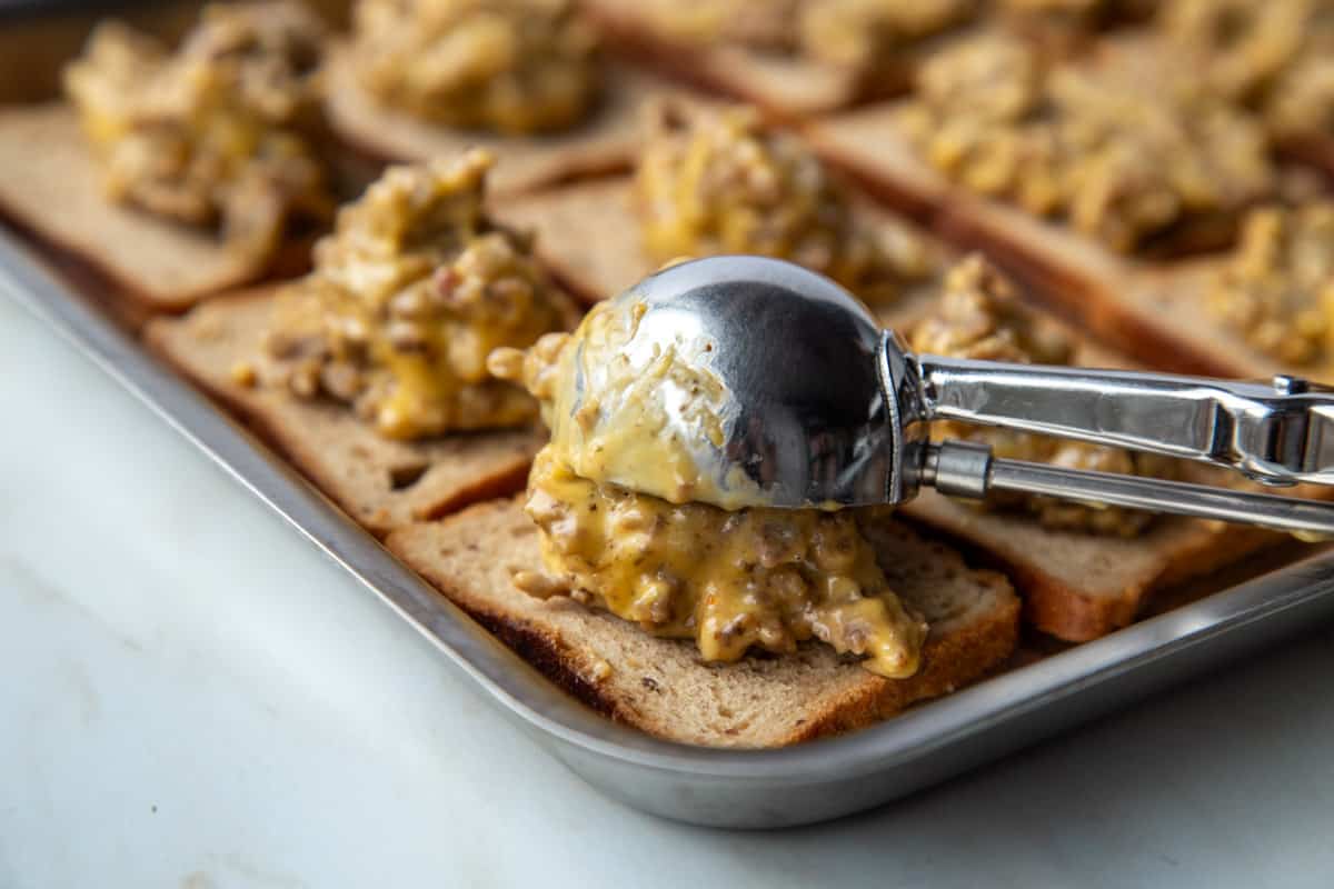 metal scoop putting meat and cheese topping on slices of rye bread on a baking sheet.